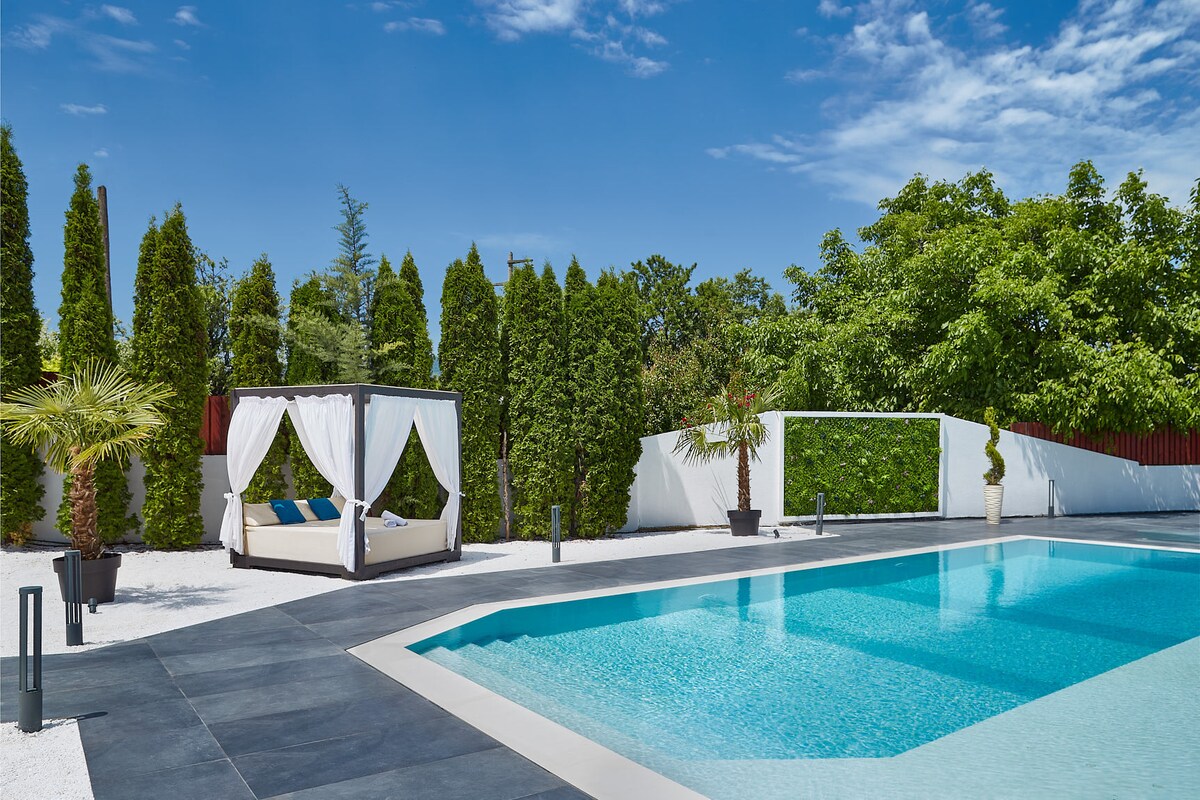 A serene outdoor pool area is surrounded by tall green hedges and decorative plants. A canopied daybed with blue cushions is positioned alongside the pool, inviting relaxation under the clear blue sky. The pool's crystal-clear water reflects the sunlight, enhancing the overall tranquil setting.