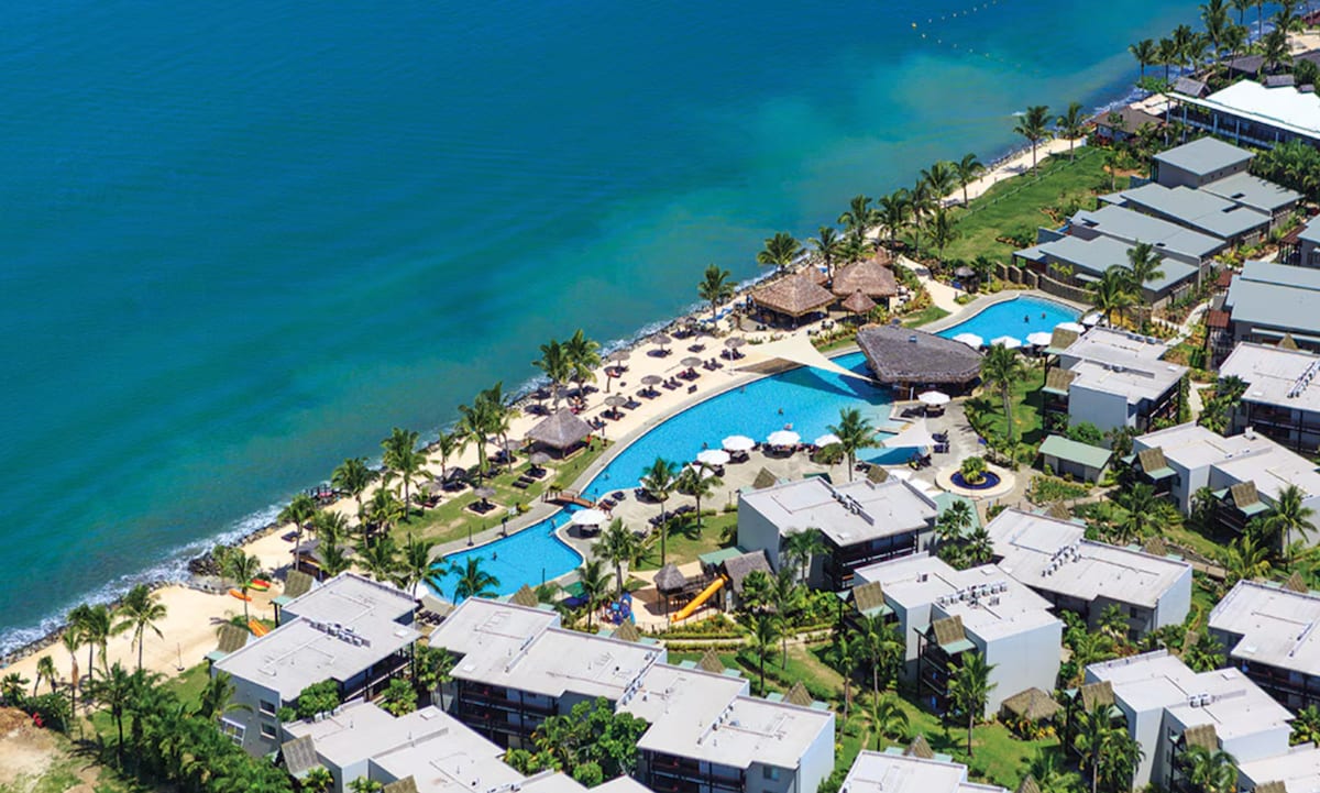 An aerial view of the resort reveals an expansive swimming pool, a sandy beach, and palm trees lining the coast. Lounge chairs and umbrellas are positioned near the pool, while the ocean's gentle waves reflect shades of blue under sunny skies.