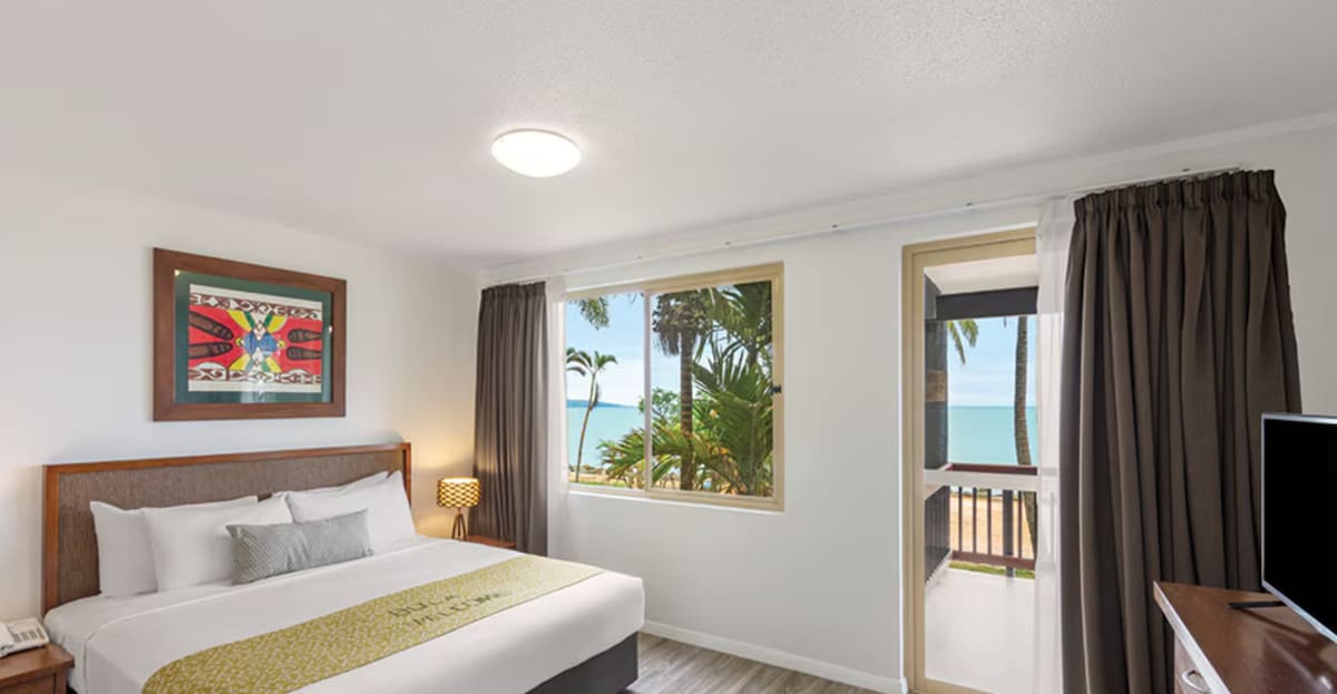 A spacious bedroom features a king bed with neutral bedding. Large windows allow natural light to fill the room while providing views of palm trees and the ocean. A side table with a lamp and a television complement the area, creating a serene and inviting space.
