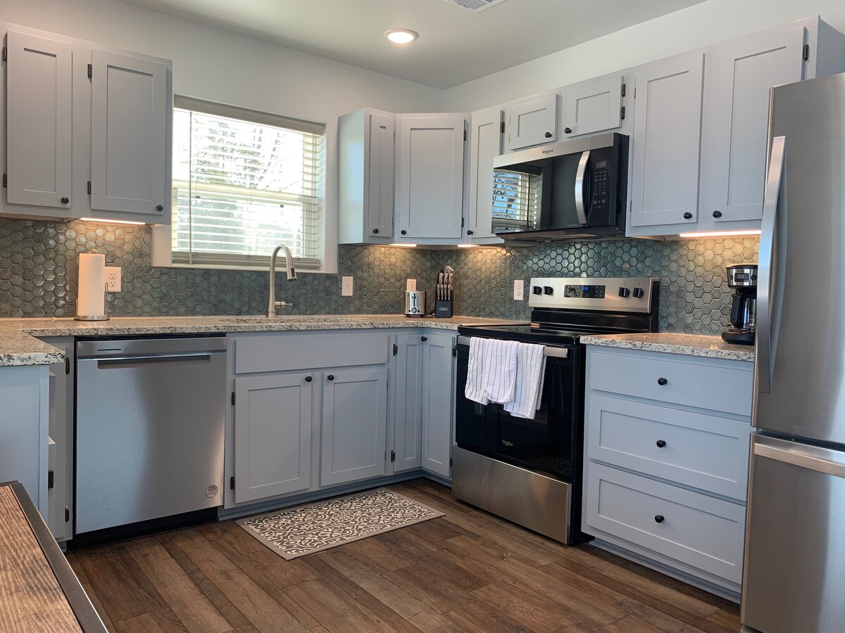 A compact kitchen features modern grey cabinetry and a range of stainless steel appliances, including a microwave and an oven. Countertops are adorned with a textured stone pattern. A window allows natural light to illuminate the space, enhancing the functional layout.