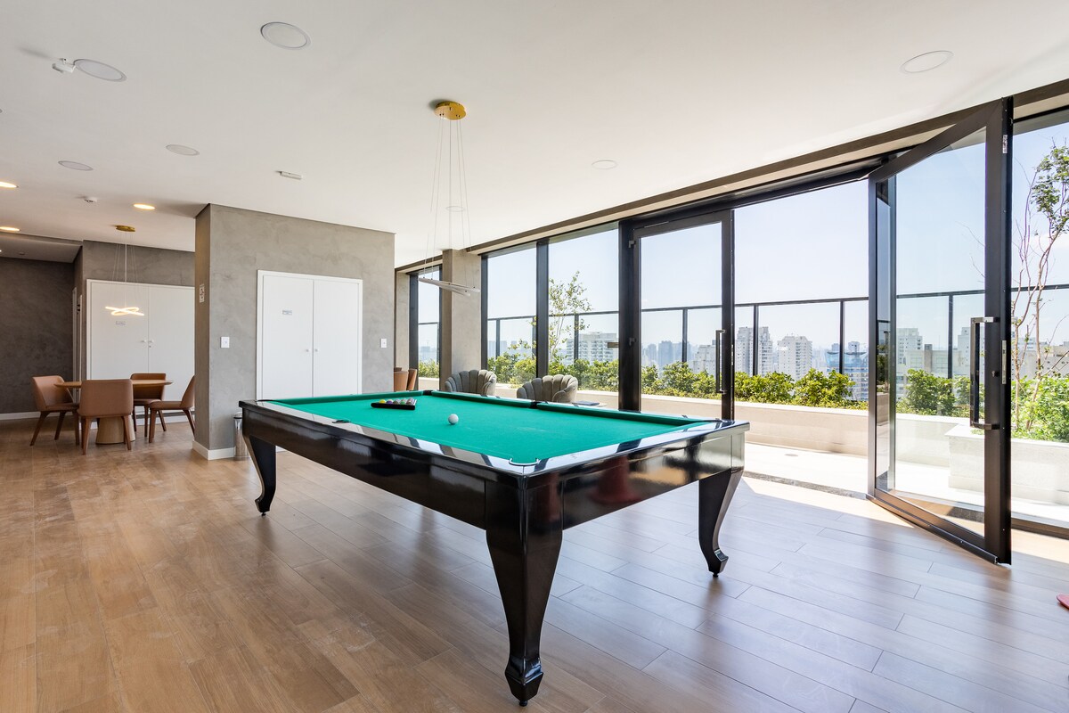 A spacious entertainment area features a green felt pool table at the center, surrounded by large windows that provide expansive views of the cityscape. Natural light floods the room, accentuating the modern decor and polished wooden floor.