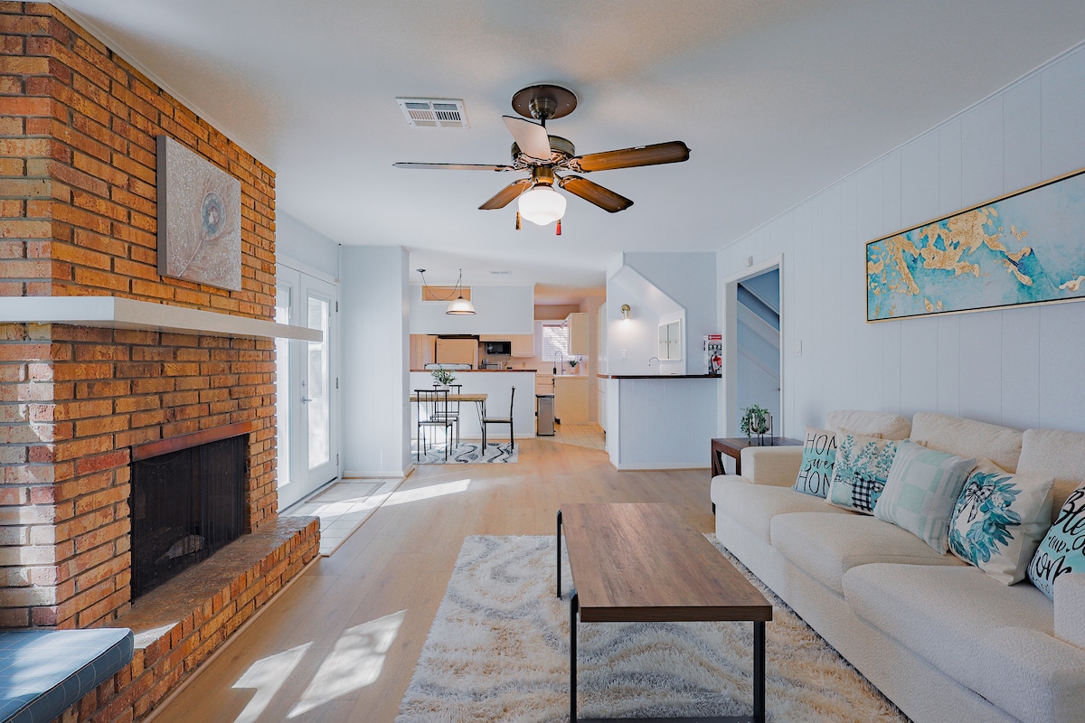 A bright living area features a cream-colored sofa adorned with decorative pillows, a coffee table, and a fireplace with a brick surround. A ceiling fan offers air circulation. Natural light fills the space through sliding glass doors leading to the kitchen area in the background.