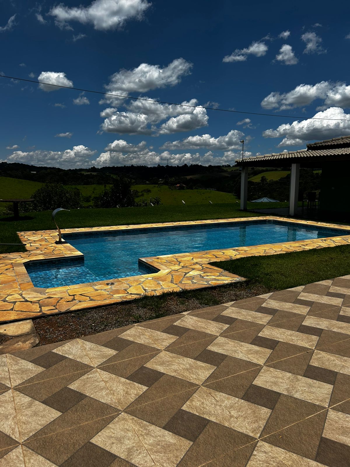 A fenced outdoor area features a clear swimming pool with a distinct shape, surrounded by a stone deck. Lush green hills are visible in the background under a bright blue sky with white clouds. A covered patio area provides shaded seating options.