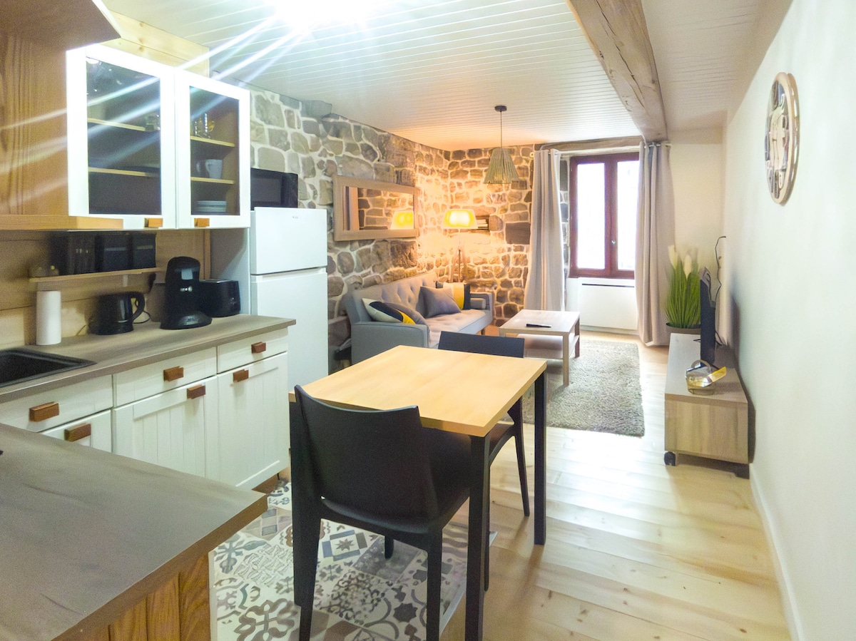 A spacious open-plan living area features a stone accent wall and wooden beams. A dining table sits beside a comfortable seating area, which is highlighted by soft lighting. Kitchen appliances, including a refrigerator and coffee maker, are visible, creating an inviting atmosphere.