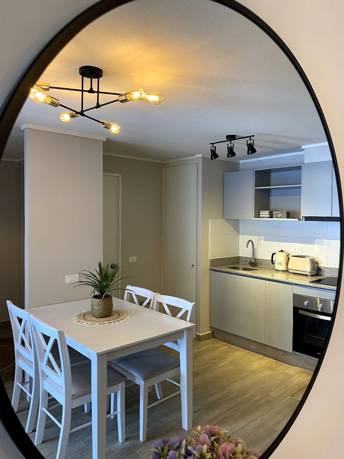 A circular mirror frames a modern kitchen and dining area. A white table with four matching chairs is set near the kitchen countertop, which features appliances and pantry items. Soft light is emitted from a ceiling fixture, enhancing the neutral color palette of the space.