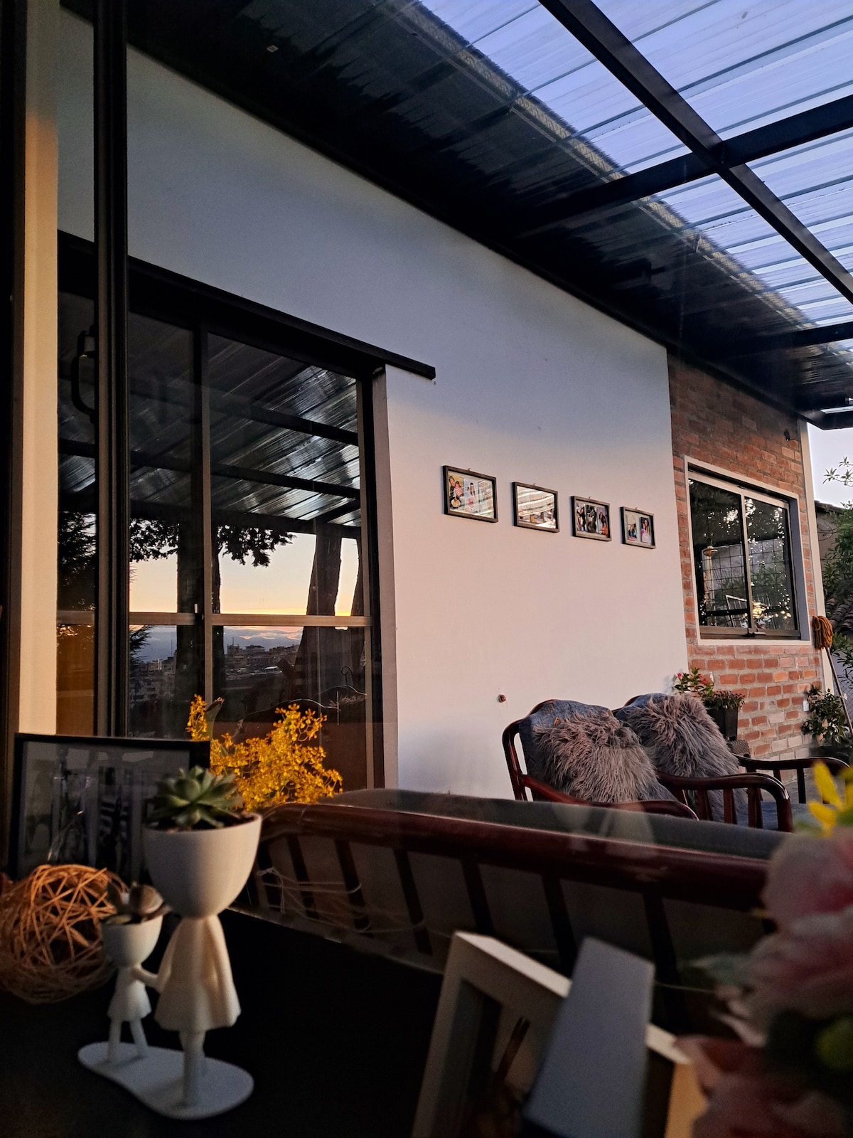 An inviting seating area is presented, featuring a wooden chair beside a glass wall. Natural sunlight illuminates the space, highlighting decor on the adjacent wall. Framed images are neatly arranged, and a small decorative figure sits on a shelf with plants adding a touch of greenery.