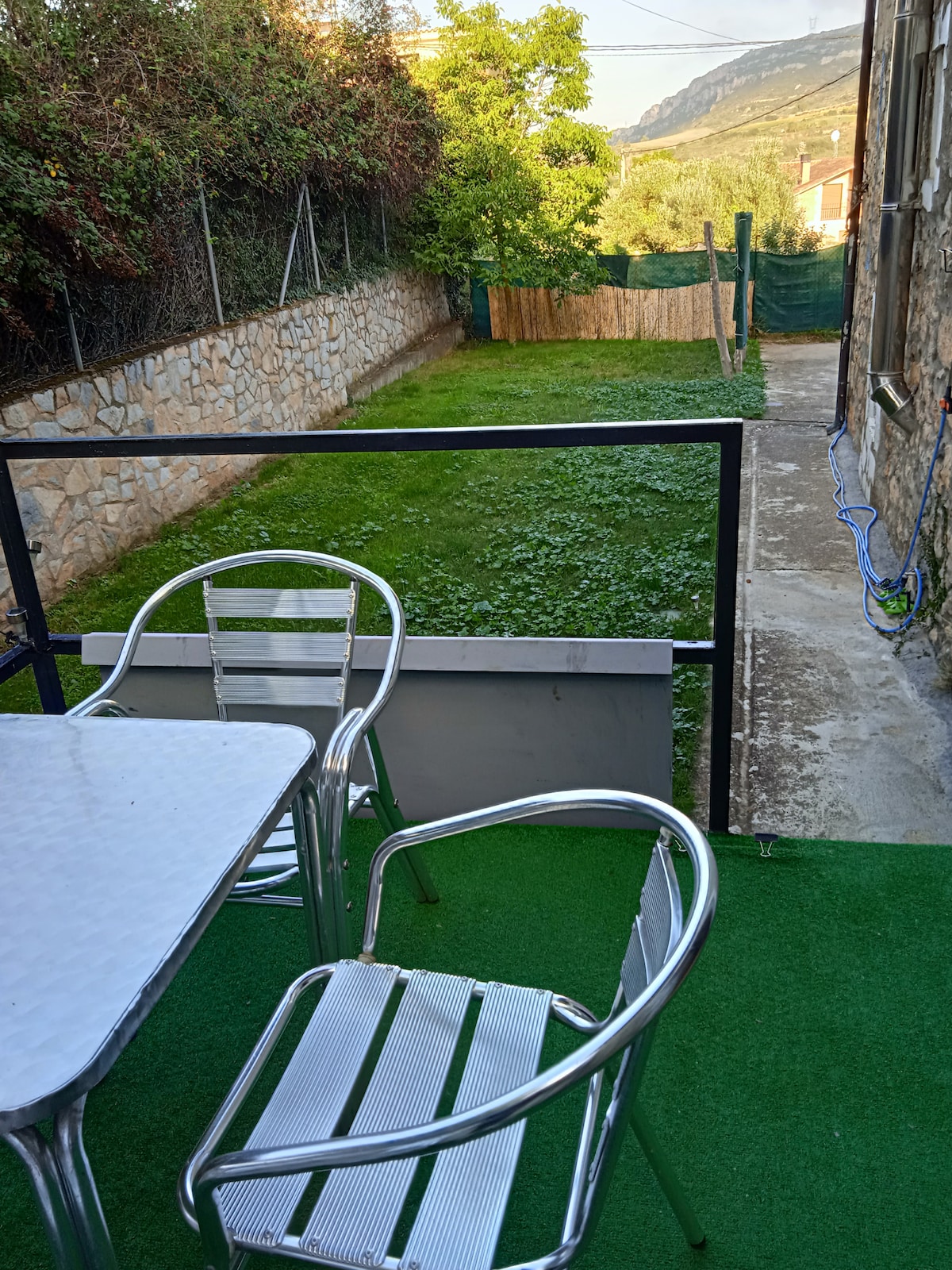 A small outdoor terrace features a table and two metal chairs, providing a space for relaxation. The view overlooks a spacious green yard, bordered by a wooden fence, with lush grass and a serene natural backdrop of trees and hills.