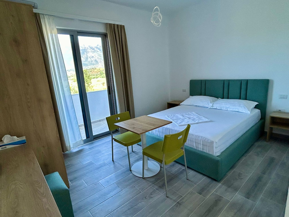A compact guest room is presented with a spacious bed covered in white linens. Two green chairs surround a small, circular table. Large glass doors open to a balcony, allowing natural light to fill the space. Neutral-toned walls and wooden accents create a modern aesthetic.