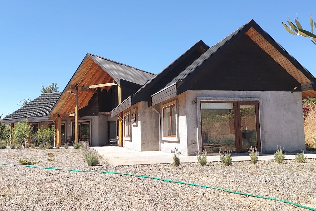 A modern house features a combination of wooden and concrete elements with a sloped roof. Large windows allow natural light to fill the interior. Surrounding greenery and a gravel area create a serene outdoor setting, enhancing the sense of tranquility in the natural landscape.