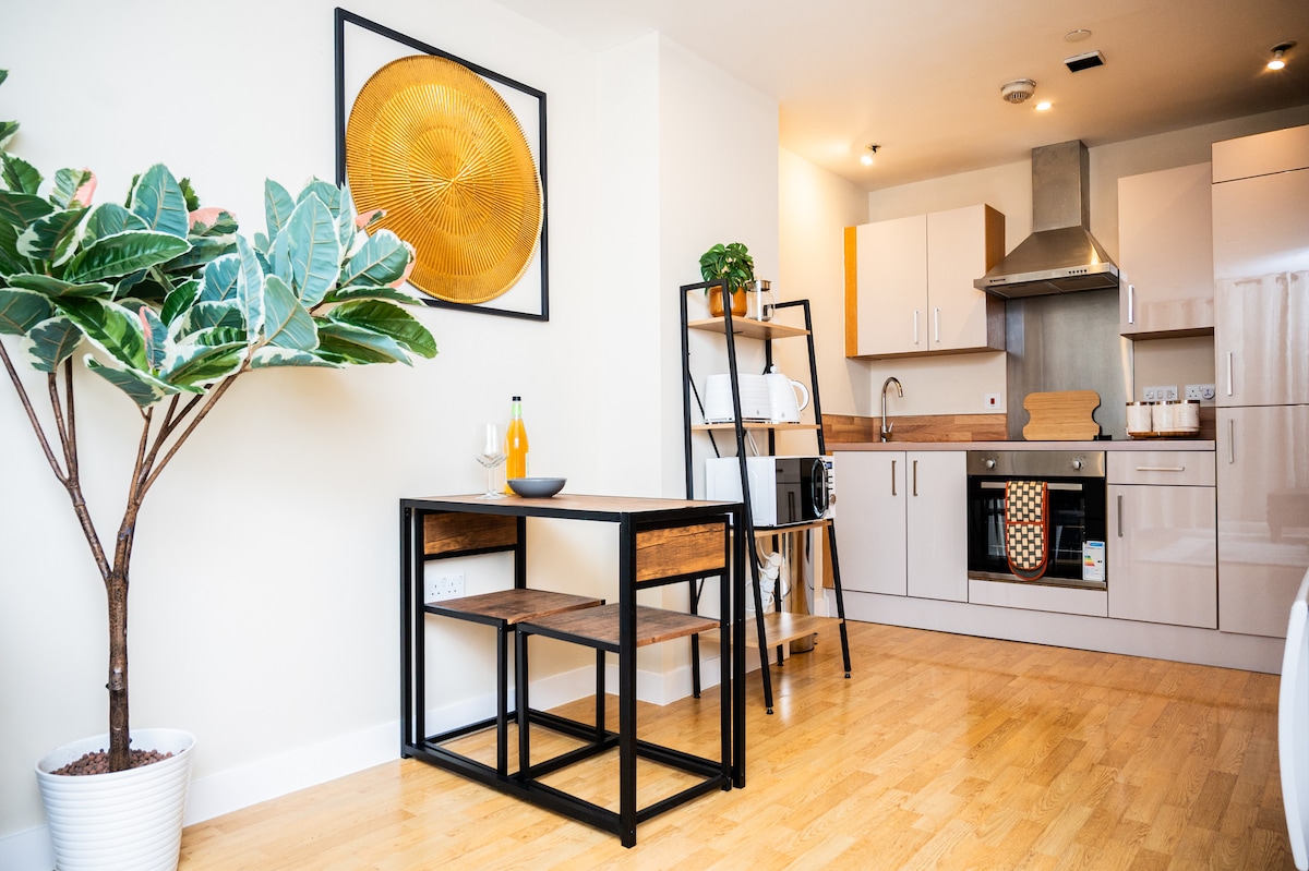 The modern kitchen area features sleek cabinetry and stainless steel appliances. A small dining table is positioned near a decorative plant. A piece of circular wall art adds a touch of style to the neutral-toned space, illuminated by soft overhead lighting.