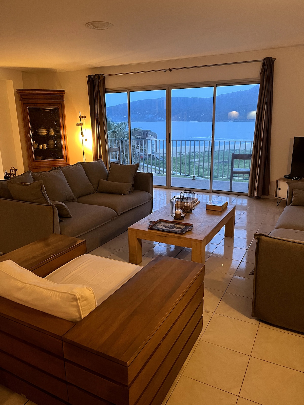 A living space features two comfortable sofas arranged around a wooden coffee table. Large glass doors provide a view of the sea, while soft lighting casts a warm glow. The decor combines modern and vintage elements, creating a cozy atmosphere.