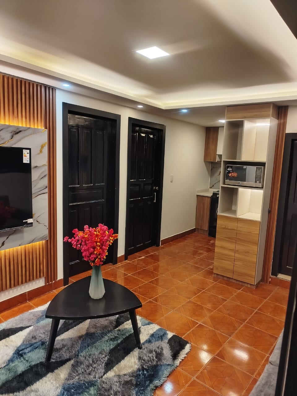 An inviting living area features a dark wooden door and a modern kitchenette in the background. A mounted television is positioned on a marbled wall, while a small black table holds a vase with colorful flowers. A textured rug adds warmth to the tile floor.