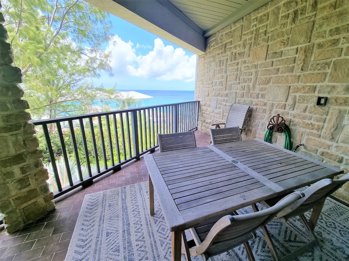A private patio showcases a wooden dining table surrounded by four chairs, set against a backdrop of ocean views. The space features a textured rug underfoot and a railing that allows glimpses of the lush greenery. Soft, natural light filters through, enhancing the tranquil setting.