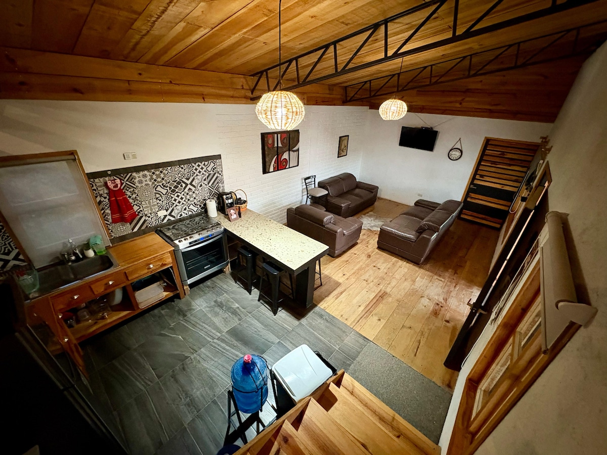 An open living area features a well-equipped kitchen and a spacious dining table. Two comfortable sofas provide seating, while a mounted television is visible on the wall. Wood-paneled ceilings and warm-toned flooring create a cozy atmosphere, enhanced by decorative light fixtures.