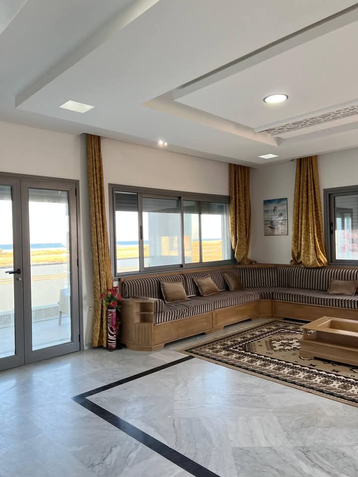 A spacious living area features a contemporary seating arrangement with striped cushions, complemented by rich golden drapes. Large windows provide abundant natural light and showcase serene views of the outdoors. A decorative area rug adds warmth, while a sleek coffee table sits at the center.
