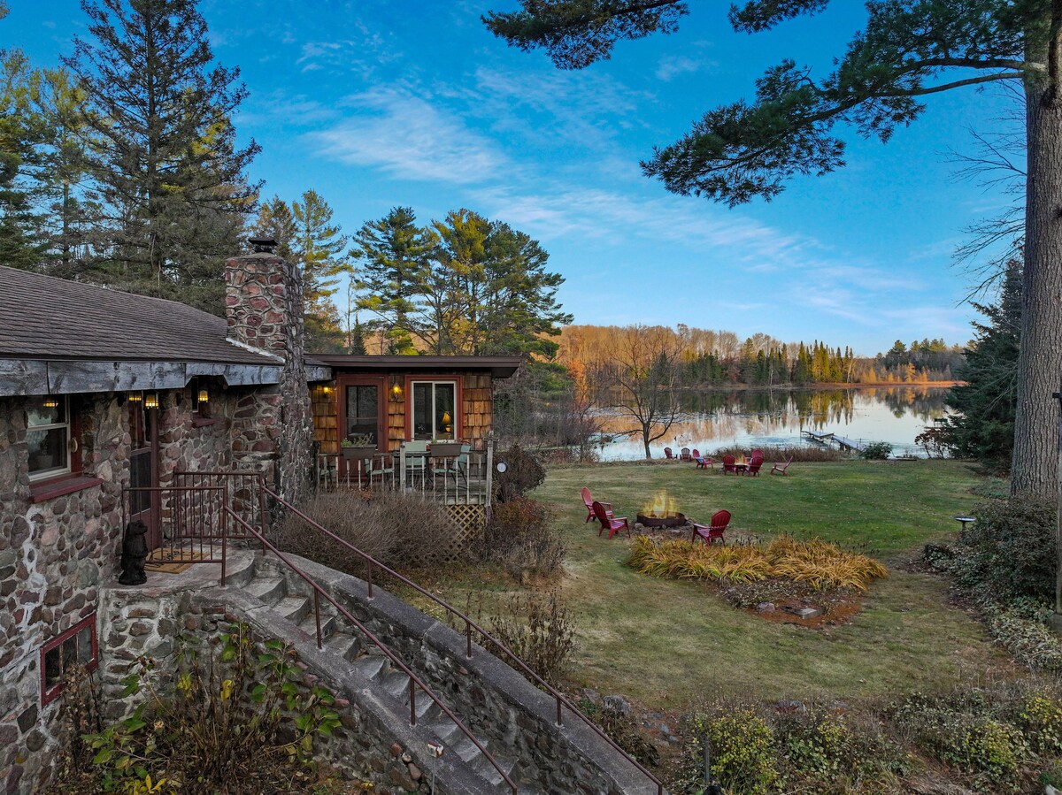 The exterior of the lodge is showcased, featuring natural stone walls and wooden accents. The well-maintained yard leads to a serene lake, reflecting the surrounding trees and sky. Comfortable red chairs are arranged near the water's edge, inviting relaxation amidst nature.