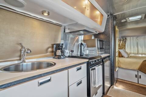 The kitchenette is equipped with a polished sink and a coffee maker, complemented by a microwave and a small stove. Sleek cabinetry above provides storage, while a warm glow from under-cabinet lights enhances the space. A window allows natural light, creating a functional cooking area.