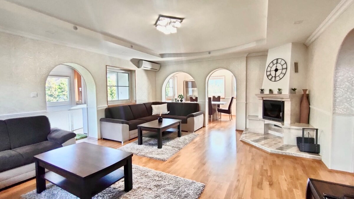A spacious living room features large windows providing ample natural light. A cozy seating arrangement with dark sofas surrounds a sleek coffee table, complemented by a modern fireplace. Arched alcoves visually enhance the room's open layout, creating a welcoming environment for gatherings.