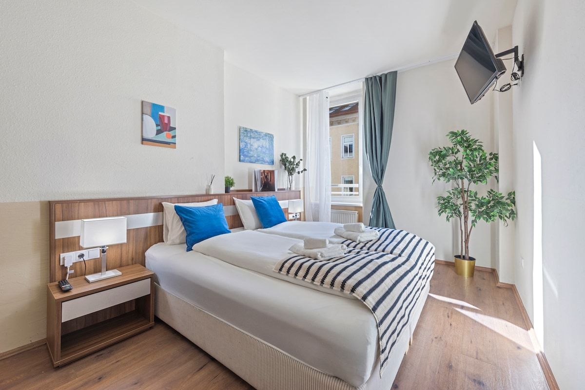 A bright bedroom is presented, featuring a spacious bed with white linens and blue accent pillows. A wall-mounted television is positioned across from the bed. Natural light filters through large windows, complemented by soft curtains, and a decorative plant adds a touch of greenery.