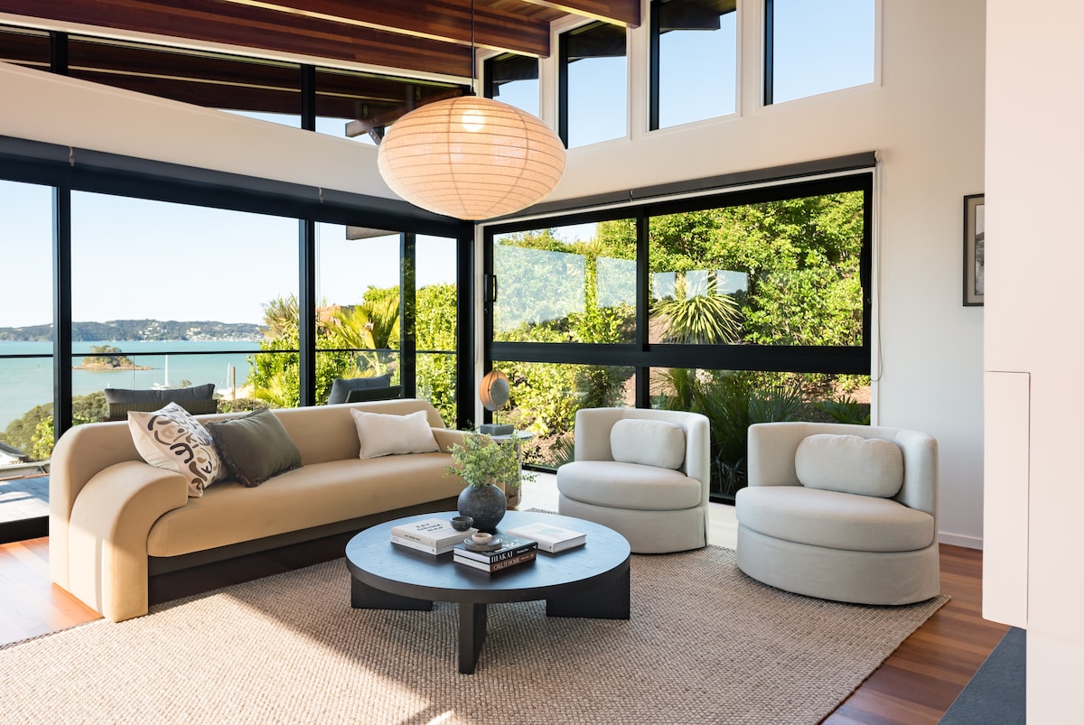 A contemporary living area features a beige sofa and two round armchairs surrounding a low black coffee table. Large windows offer views of the lush greenery and water beyond, while natural light fills the space. A pendant light hangs above, adding a soft glow.