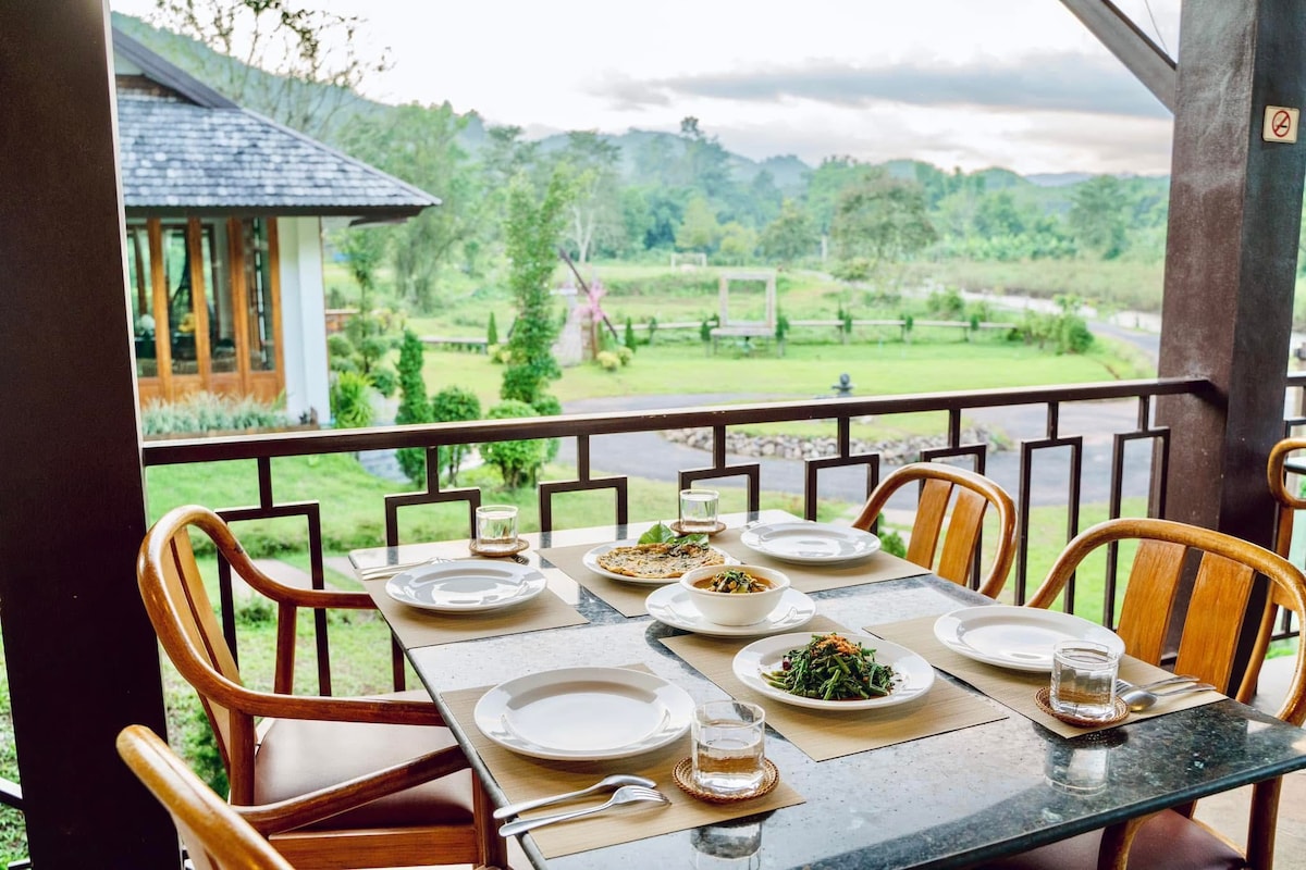 A dining table is set on a covered balcony, featuring four wooden chairs around it. Plates of food are arranged alongside glasses, with a scenic view of lush greenery and distant hills visible in the background.
