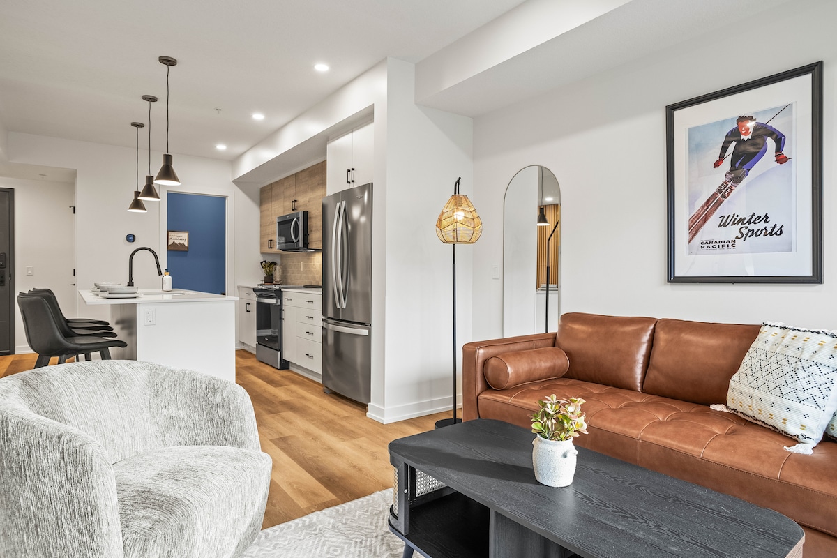 An open living space showcases modern furnishings, including a sleek leather couch and a textured armchair. A coffee table is positioned in front, while a fully equipped kitchen with stainless steel appliances and dark cabinetry is visible in the background. Bright, natural light fills the area.