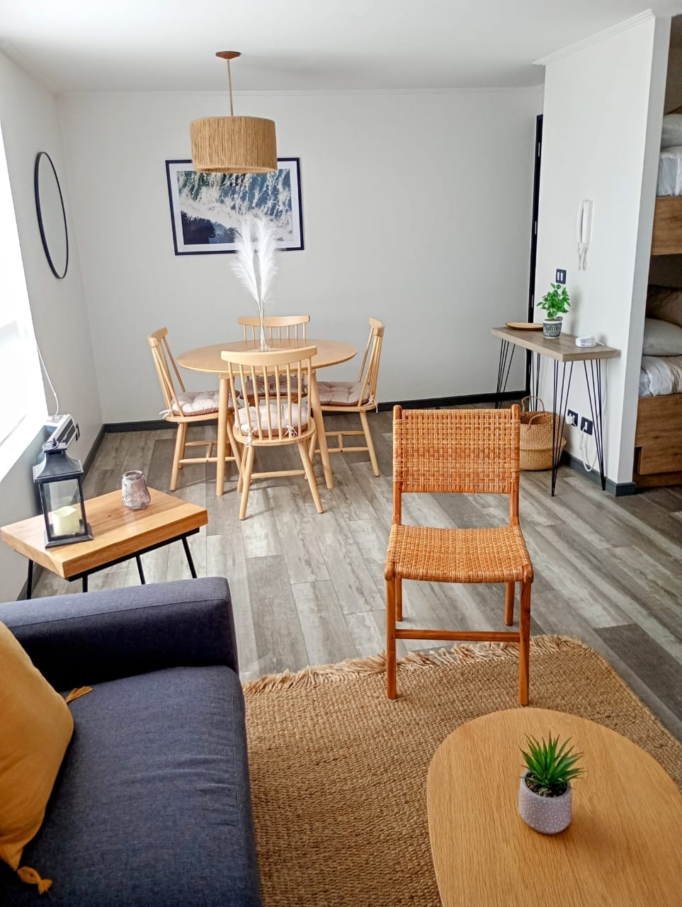 A modern living space features a cozy corner sofa and a round dining table surrounded by four wooden chairs. A woven pendant light hangs above the table, and a decorative plant is positioned on a side table. Natural light enters through a large window, illuminating the room.