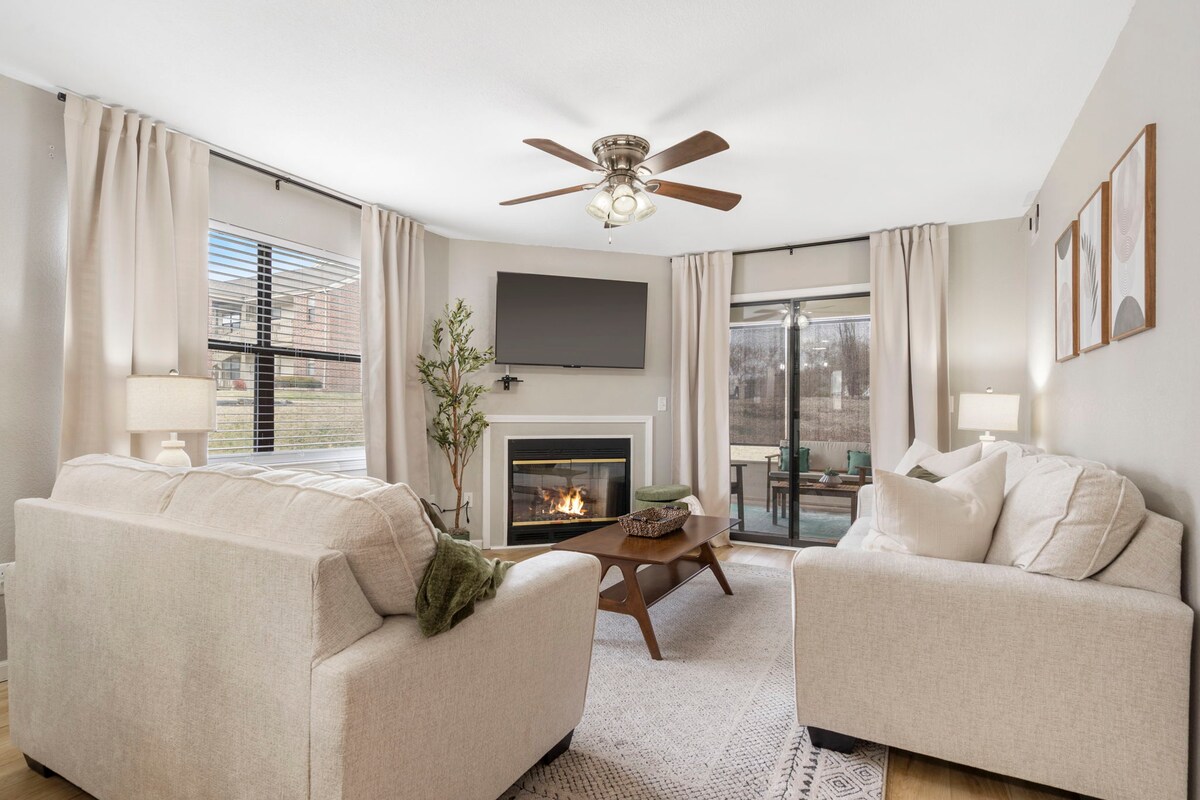 The living room features two inviting sofas arranged around a central coffee table, with a mounted TV for entertainment. A ceiling fan and natural light from large windows create a bright atmosphere. A sliding door leads to a patio area, enhancing the space's openness.