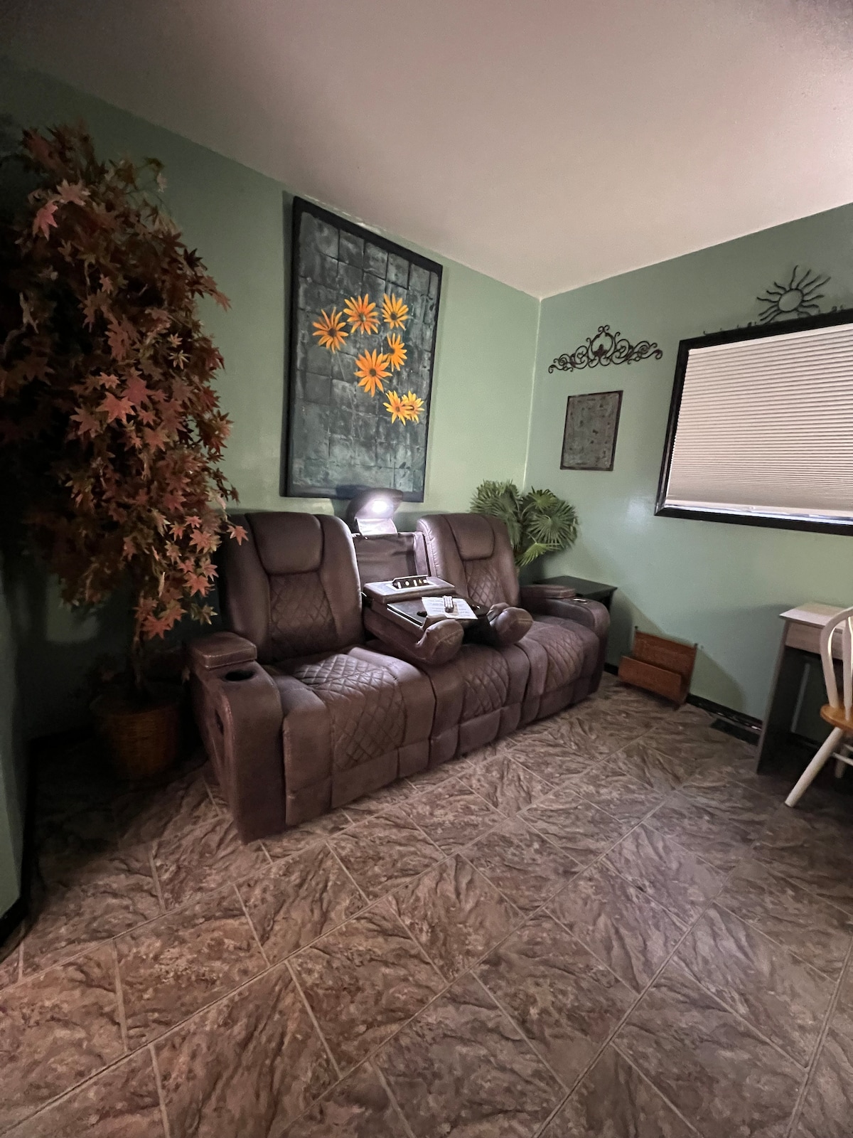 A comfortable recliner sofa is positioned in a cozy corner of the living space, accompanied by a potted plant and decorative wall art depicting flowers. Natural light filters through a partially closed window, providing a soft, serene atmosphere.