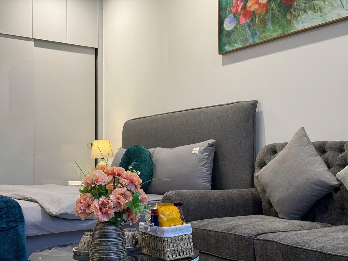 A cozy living area is presented, featuring a grey sofa adorned with patterned cushions. A small table holds a bouquet of pink flowers and snacks. A soft bed is partly visible in the background, complemented by a decorative painting above the sofa.