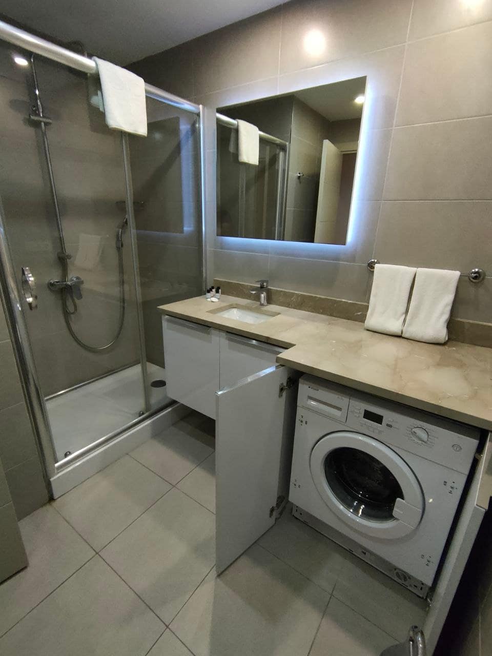 A modern bathroom is equipped with a glass-enclosed shower featuring a rainfall showerhead. A sink with a smooth countertop provides space for toiletries. A washing machine is integrated into the cabinetry, and white towels are neatly displayed on the counter.
