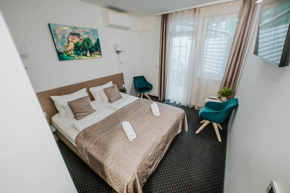 A room features a spacious bed with neutral bedding and decorative pillows. Two teal chairs are positioned by the window, complemented by a small plant. Natural light filters through sheer curtains, and a piece of wall art adds a touch of color.