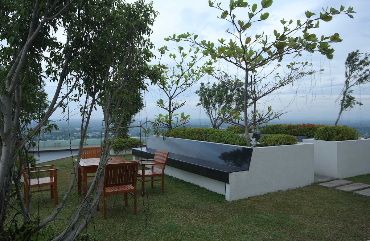 An outdoor communal area showcases wooden furniture arranged around a circular table, surrounded by lush greenery. Delicate trees rise above, providing shade and a natural ambiance. The space offers panoramic views, with soft grass underfoot and planters lining the perimeter.