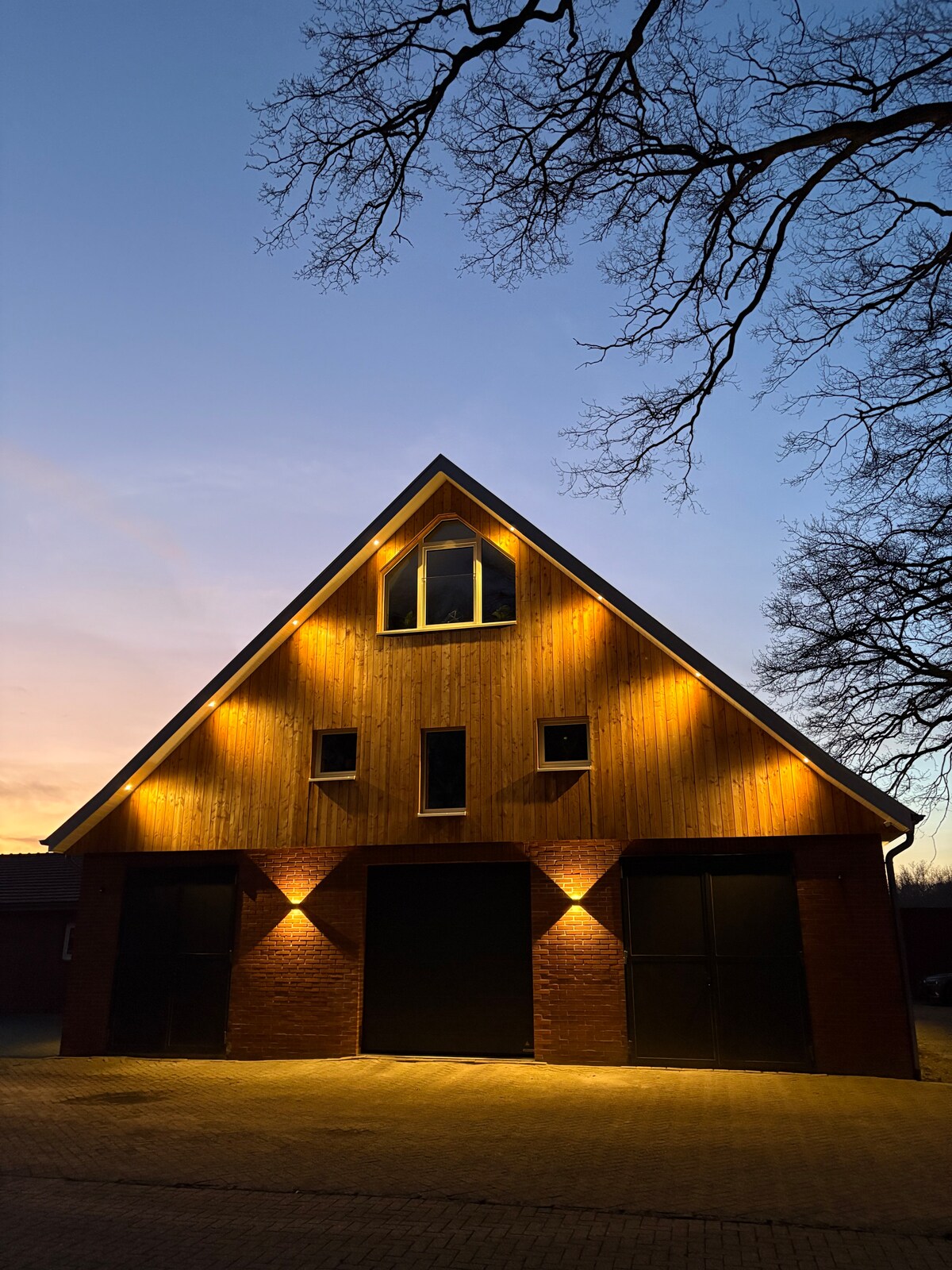 The rustic barn structure features a gabled roof with soft lighting highlighting the wooden facade. Large, dark doors serve as the main entrance, flanked by smaller windows that allow for natural light. The setting sun creates a gentle glow in the sky.