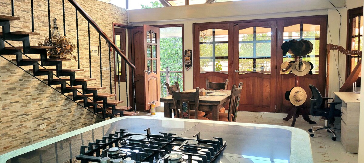 The open kitchen features a modern cooktop with multiple burners, complemented by a rustic dining area with wooden chairs and a table. Large glass doors provide natural light and a view of the outdoor space, while a staircase leads to an upper level.
