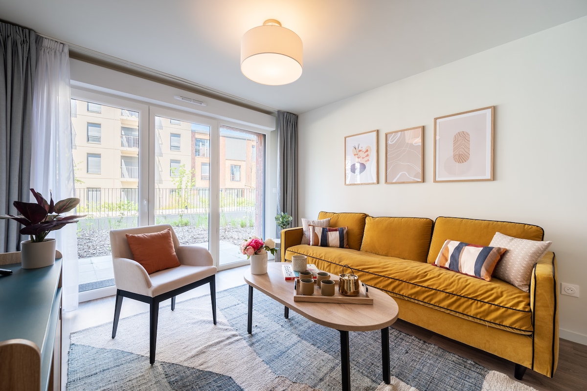 A bright and inviting living area is presented, featuring a yellow sofa adorned with decorative cushions. A small wooden coffee table is positioned in front, accompanied by a comfortable chair. Large sliding glass doors provide access to an outdoor space, enhancing the room's natural light.