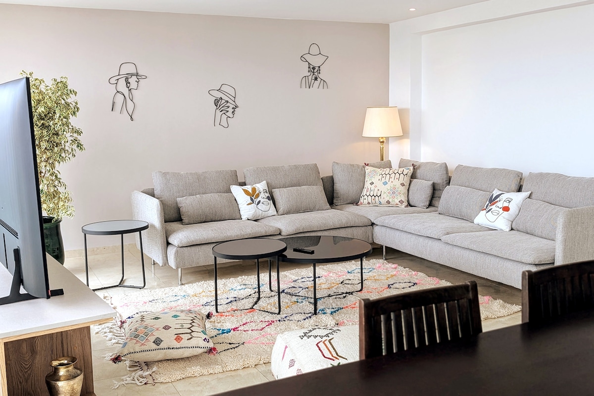 A spacious living area is featured, furnished with a large sectional sofa adorned with colorful cushions. Two black side tables are positioned next to the sofa, and an area rug adds texture beneath. Minimalist wall art of human figures is displayed on a light-colored wall, and a floor lamp provides soft lighting.