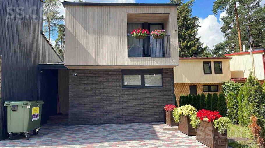 A modern home exterior is showcased, featuring a light-colored upper level with flower boxes, a balcony, and dark brick lower walls. Vibrant flowers and well-maintained plants are arranged in planters along a paved pathway, contributing to the inviting appearance of the property.