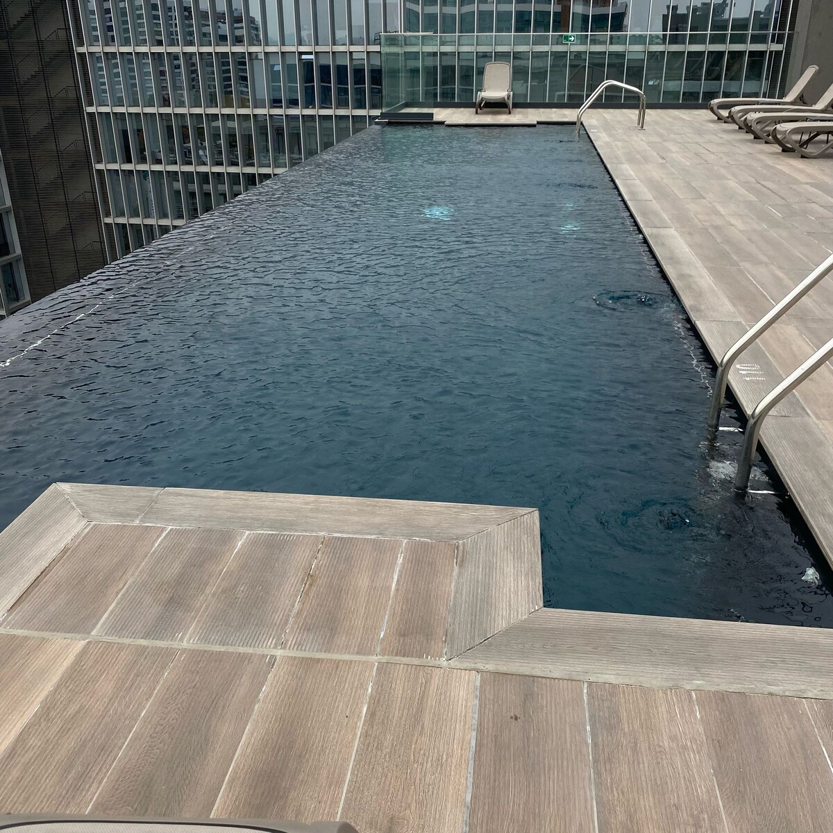An infinity pool is positioned on a rooftop, featuring smooth wooden decking around its edges. The water appears calm and reflects the surrounding glass buildings. Lounge chairs are arranged nearby, providing a relaxing atmosphere with expansive views of the city skyline.