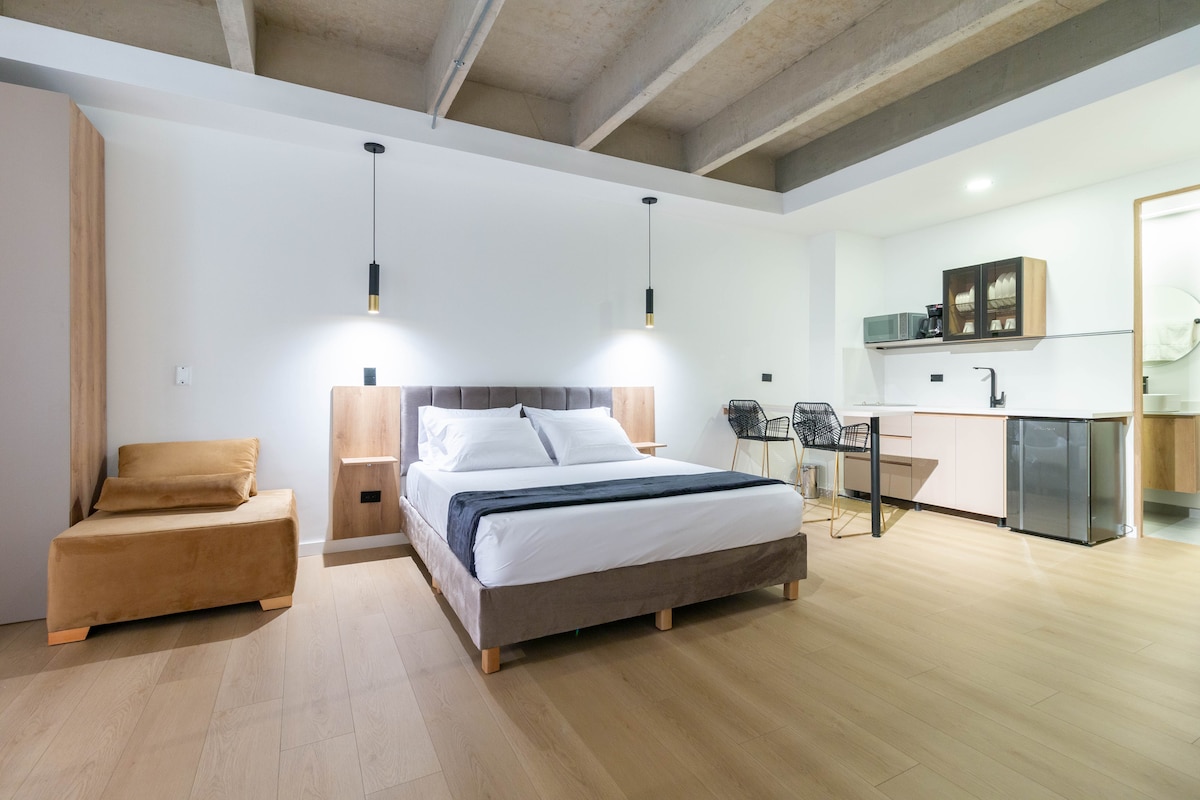 A modern, open-plan loft features a spacious bed with neutral linens. A cozy seating area is complemented by a kitchenette with high-quality appliances in the background. Soft lighting from pendant fixtures enhances the minimalist design and creates a welcoming atmosphere.
