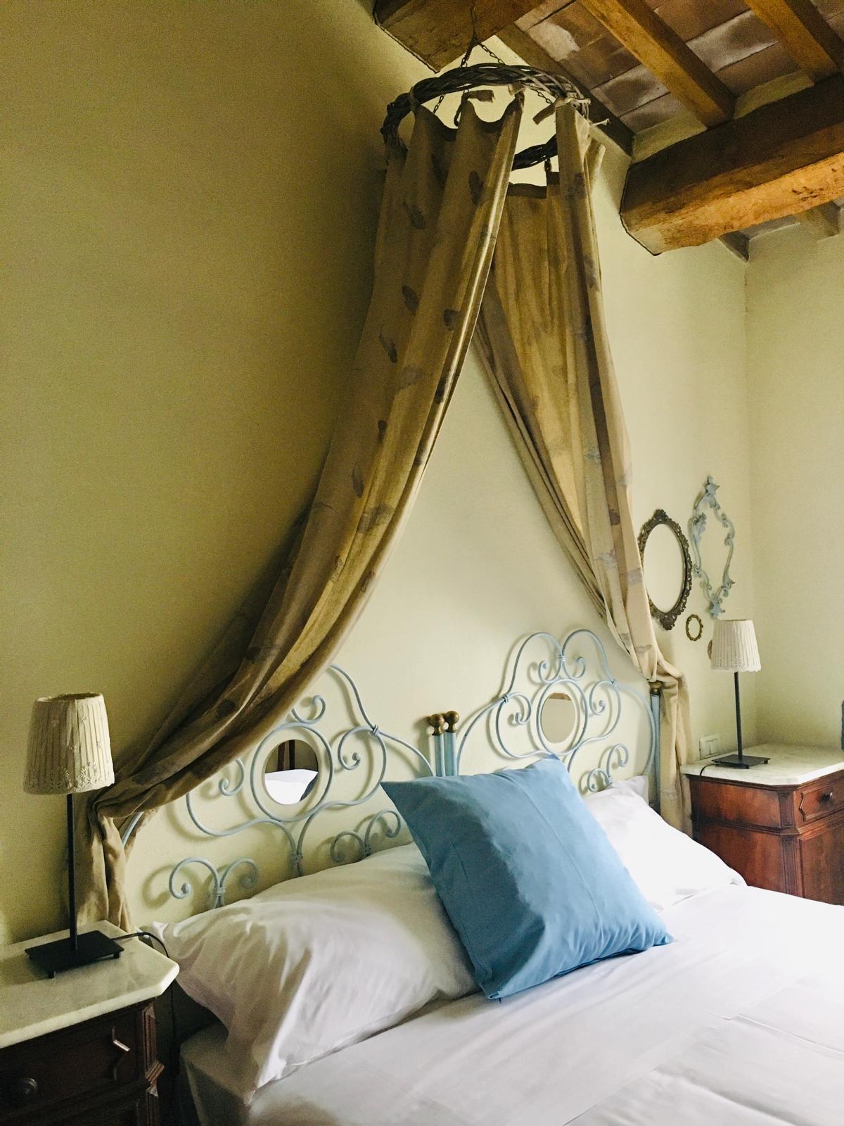 A bedroom features a wrought iron headboard adorned with fabric draped elegantly overhead. A soft blue cushion complements the white bedding. Two bedside lamps provide gentle illumination, while decorative mirrors are displayed on the walls, contributing to a serene atmosphere.