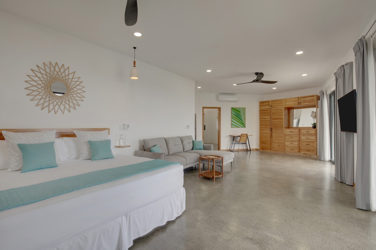 A spacious hotel room is featured, showcasing a large King bed with soft linens and decorative pillows. A comfortable sofa sits adjacent, with a small coffee table in front. Natural light fills the room through sliding glass doors, which lead to the private patio.