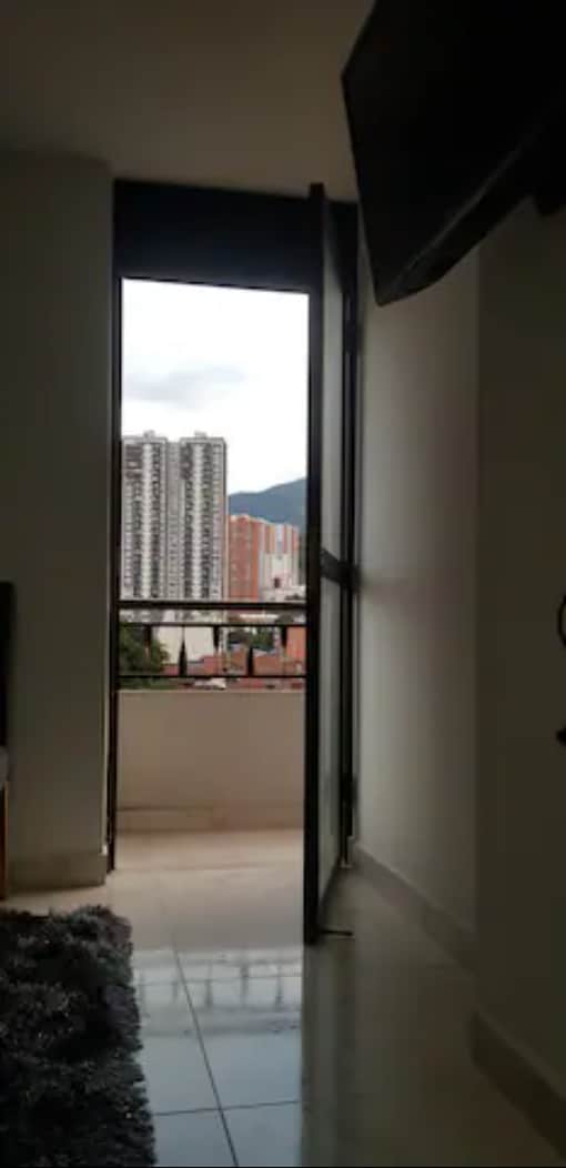 A corner of the room is captured featuring an open door leading to a balcony, allowing for views of surrounding buildings and mountains in the distance. The space is bright, with natural light entering through the doorway, illuminating the contemporary interior.