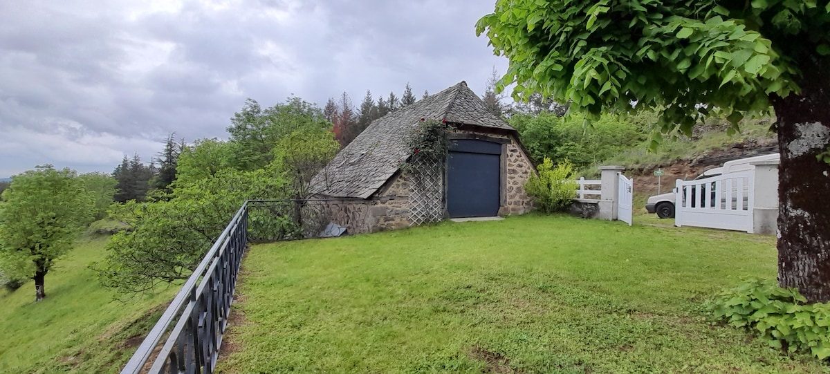 A quaint stone building with a sloped roof is set within a grassy area, surrounded by lush trees. A dark door is prominently displayed, and a railing runs along the grassy path leading to the structure. The peaceful landscape offers a serene backdrop.