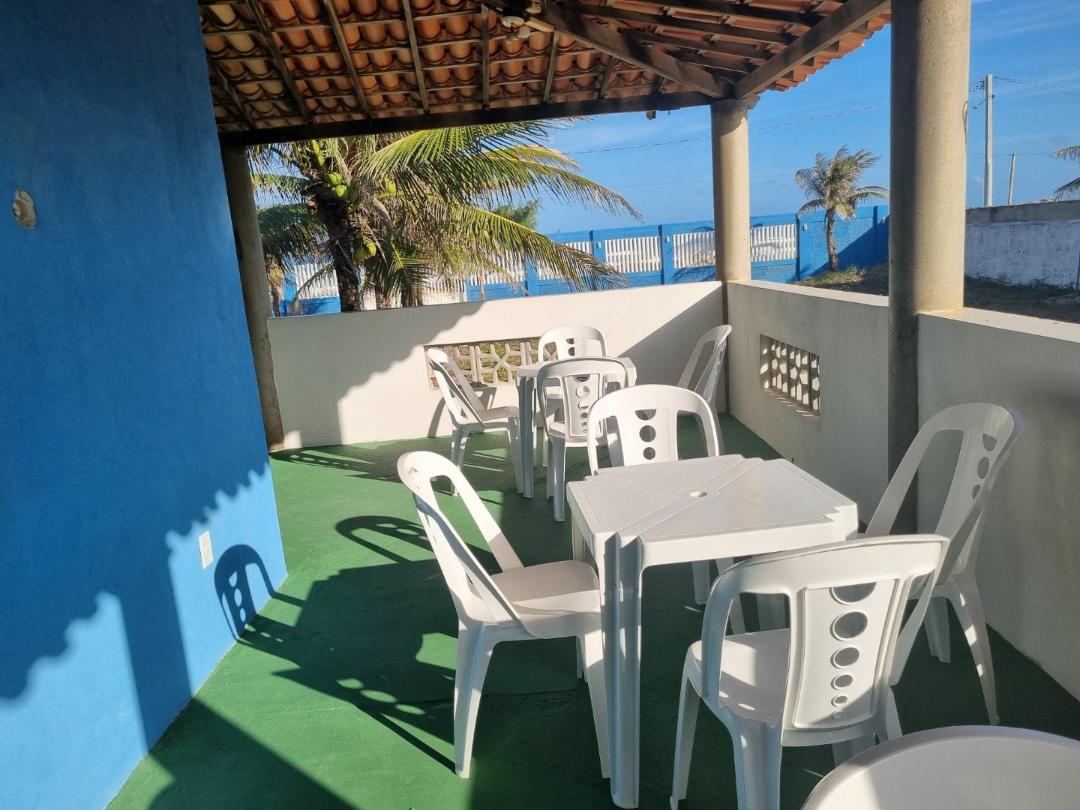An outdoor seating area features white plastic tables and chairs arranged on a green floor. A blue wall provides a vibrant backdrop, while palm trees and the ocean are visible in the distance, creating a relaxed atmosphere under a partially covered roof.