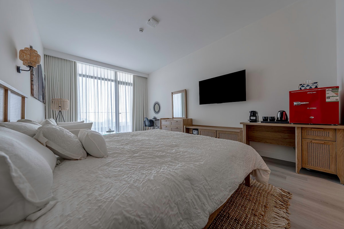 A spacious bedroom features a large bed with white bedding and decorative pillows. Natural light floods the room through floor-to-ceiling windows, while a flat-screen television is mounted on the wall. A wooden dresser and a vibrant red mini fridge are featured in the room.