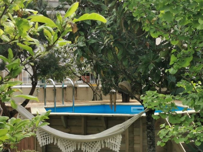 A serene outdoor area is framed by lush greenery, featuring a clear blue above-ground pool partially visible in the background. A hammock is draped between wooden posts, inviting relaxation in a tranquil setting.