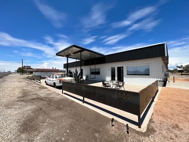A modern exterior view of The Coupe Cowboy micro-hotel is presented, featuring a covered patio with a dining table and chairs. An aged vintage car is parked nearby, surrounded by desert landscape. The building's clean lines are complemented by a clear blue sky and wispy clouds.