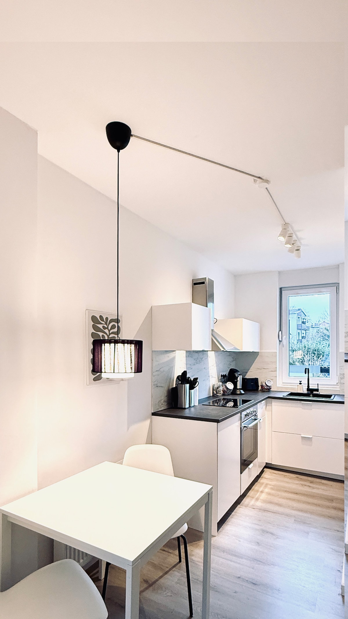 A compact kitchen area is shown, featuring modern white cabinetry and a sleek countertop. A small table with two white chairs is positioned beside a window, allowing natural light to enter. A stylish pendant light hangs from the ceiling, illuminating the space.