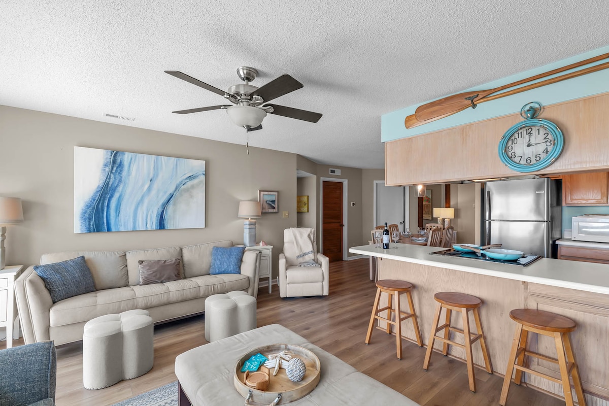 The spacious living area features a beige sofa paired with two ottomans and a decorative coffee table. A ceiling fan hangs above, and natural light floods in from the nearby windows. A large painting in soothing blue tones adorns the wall, complementing the relaxed ambiance.